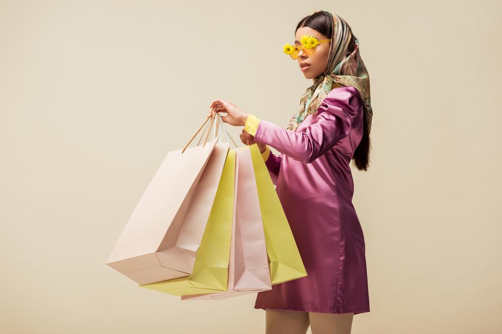 stylisches afrikanisch-amerikanisches Mädchen in Sonnenbrille mit Blumen, Kopftuch und Kleid mit Einkaufstaschen in Beige  - Foto, Bild