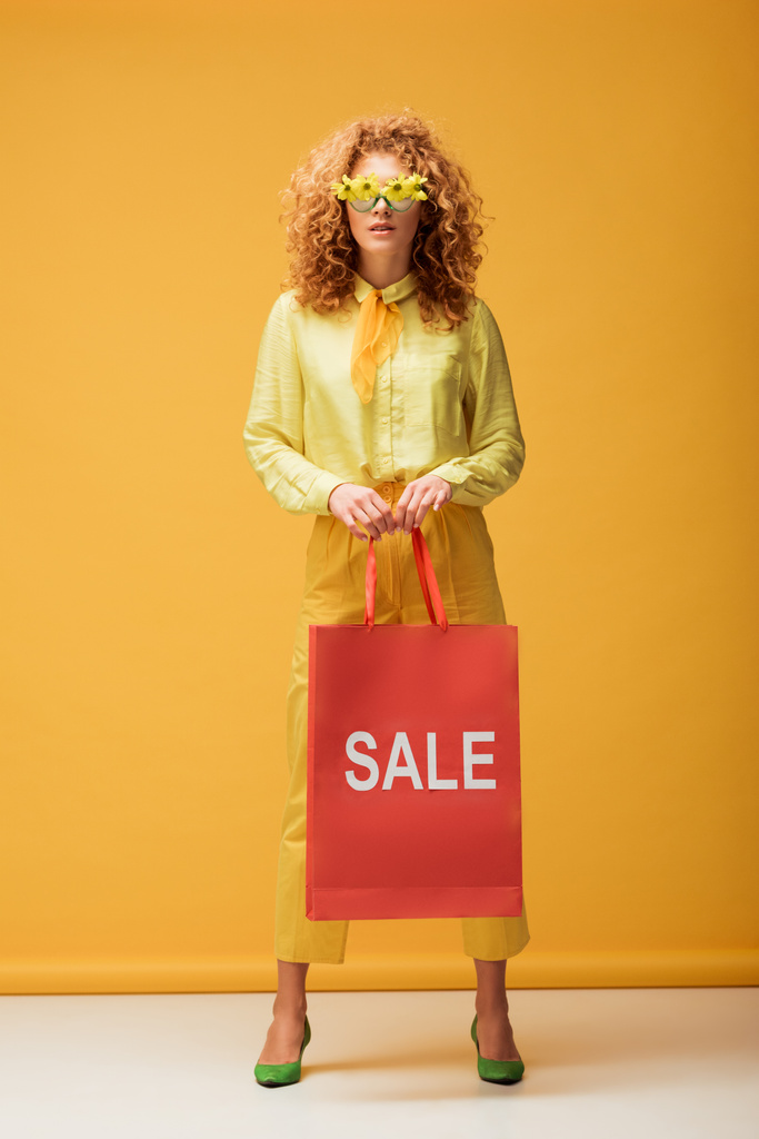 stylische rothaarige Frau mit Sonnenbrille und Blumen, die Einkaufstasche mit Verkaufsaufdruck auf gelb hält  - Foto, Bild