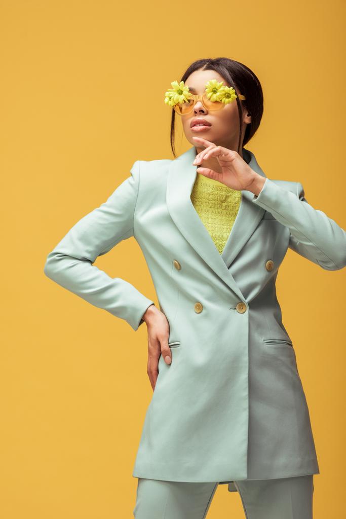 όμορφη Αφροαμερικανή με κοστούμι και γυαλιά ηλίου με λουλούδια που ποζάρουν απομονωμένα στο κίτρινο  - Φωτογραφία, εικόνα