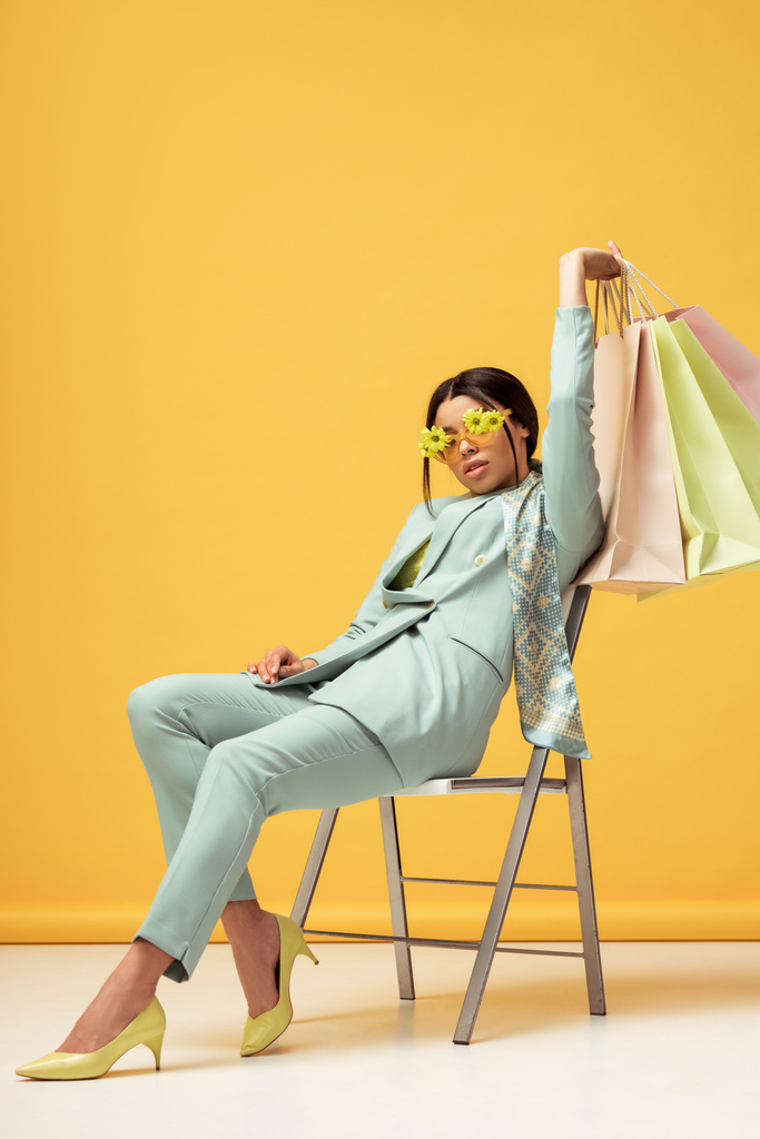 donna afroamericana alla moda in abito, sciarpa e occhiali da sole con fiori che tengono le borse della spesa mentre si siede sul giallo
  - Foto, immagini