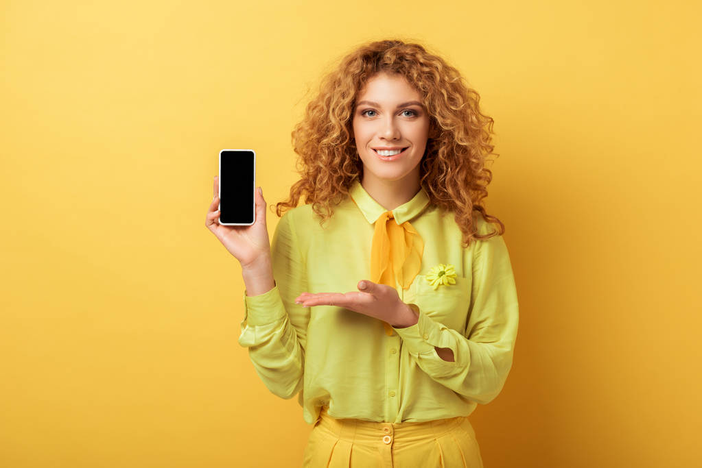 ευτυχισμένη κοκκινομάλλα γυναίκα δείχνει με το χέρι στο smartphone με λευκή οθόνη που απομονώνονται σε κίτρινο  - Φωτογραφία, εικόνα