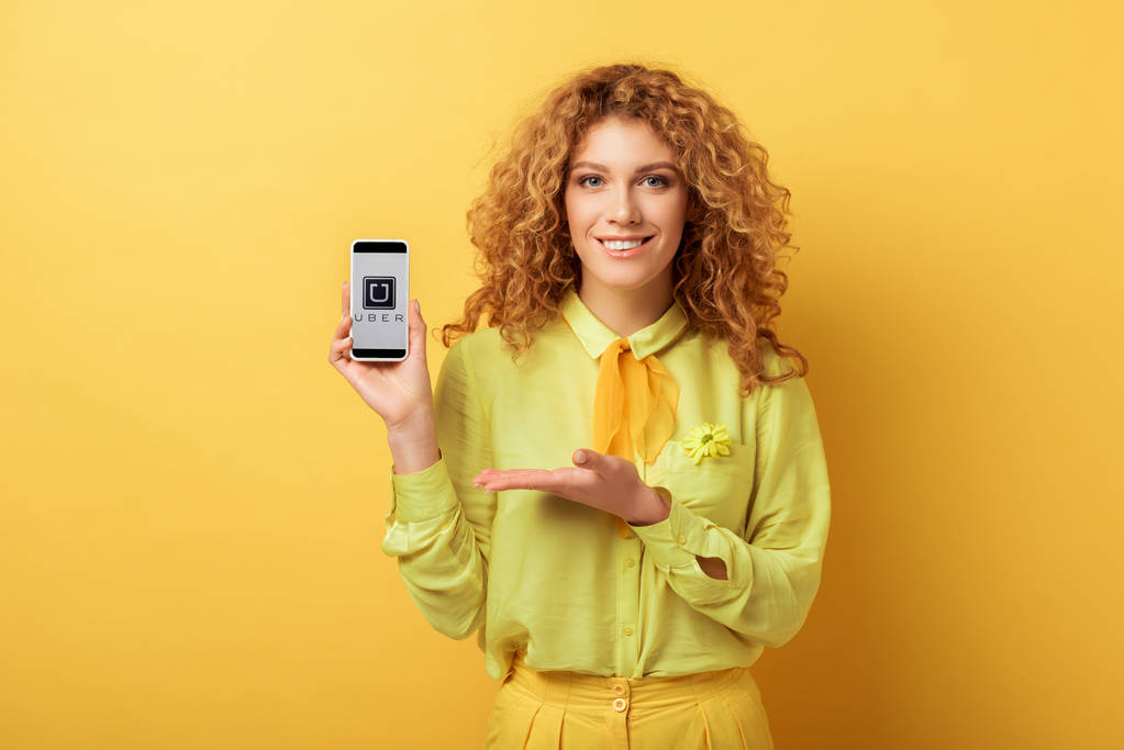 KYIV, UKRAINE - FEBRUARY 4, 2020: happy redhead woman pointing with hand at smartphone with uber app on screen isolated on yellow  - Photo, Image