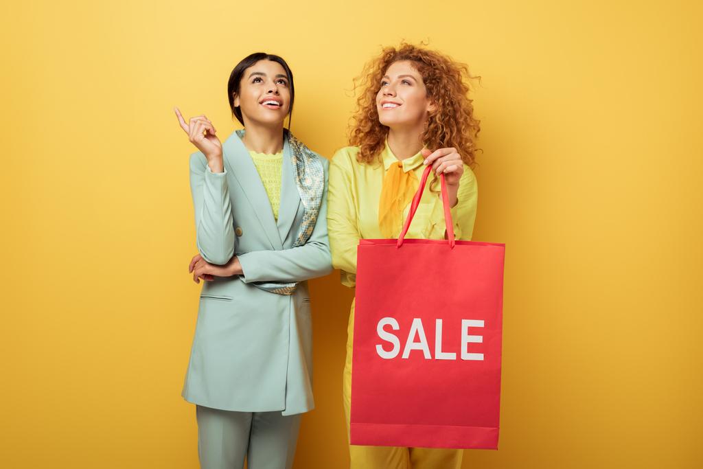 glücklich rothaarige Mädchen mit Einkaufstasche mit Verkauf Schriftzug in der Nähe lächelnde afrikanisch-amerikanische Frau zeigt mit dem Finger auf gelb  - Foto, Bild