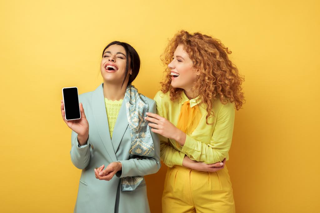 feliz afroamericano chica sosteniendo teléfono inteligente pantalla en blanco y riendo con pelirroja en amarillo
  - Foto, Imagen