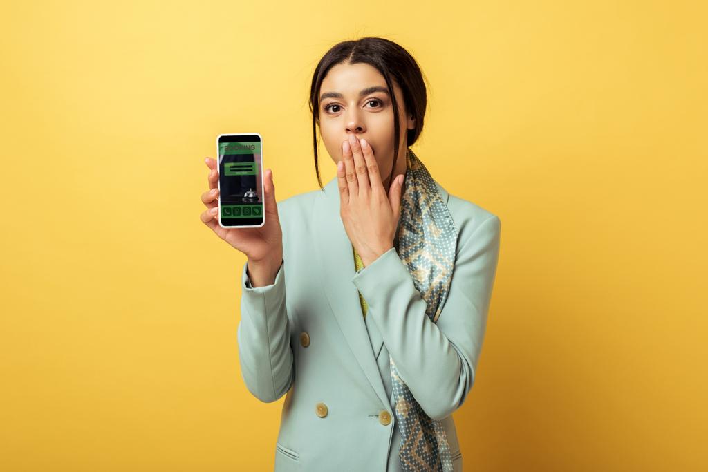 geschokt Afrikaans Amerikaans meisje bedekking gezicht en het houden van smartphone met boeking app op geel  - Foto, afbeelding