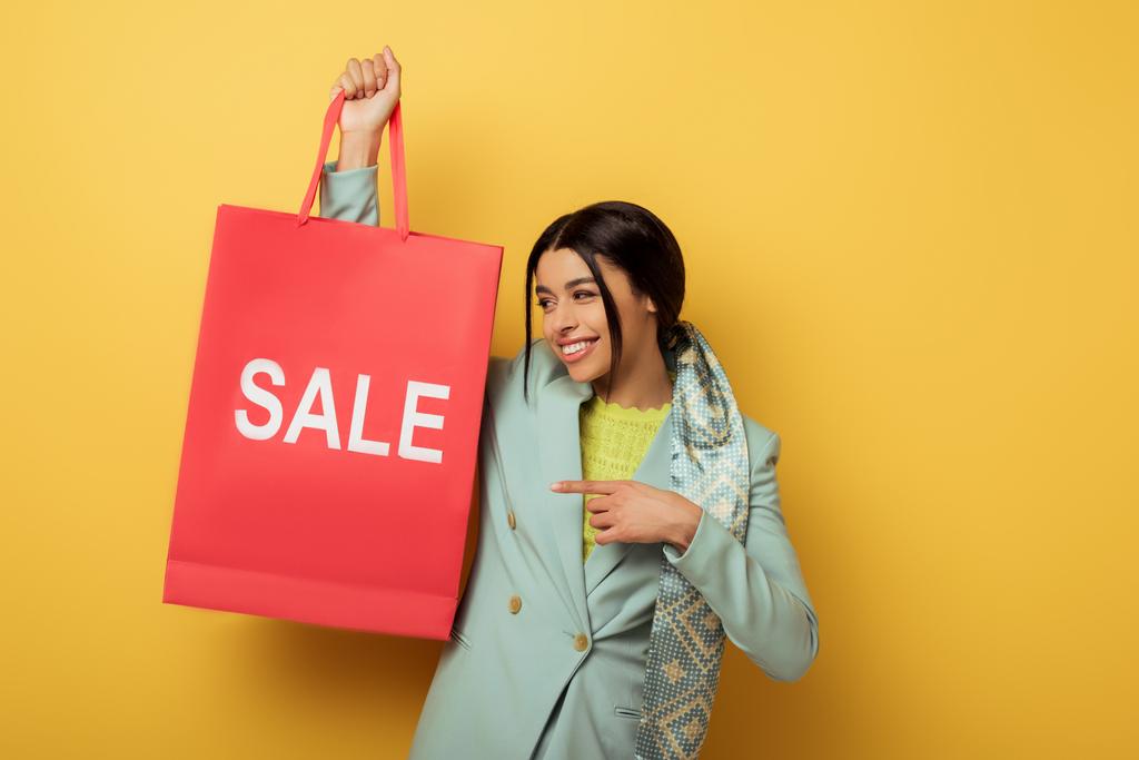 fröhliche afrikanisch-amerikanische Mädchen zeigt mit dem Finger auf Einkaufstasche mit Verkauf Schriftzug auf gelb  - Foto, Bild