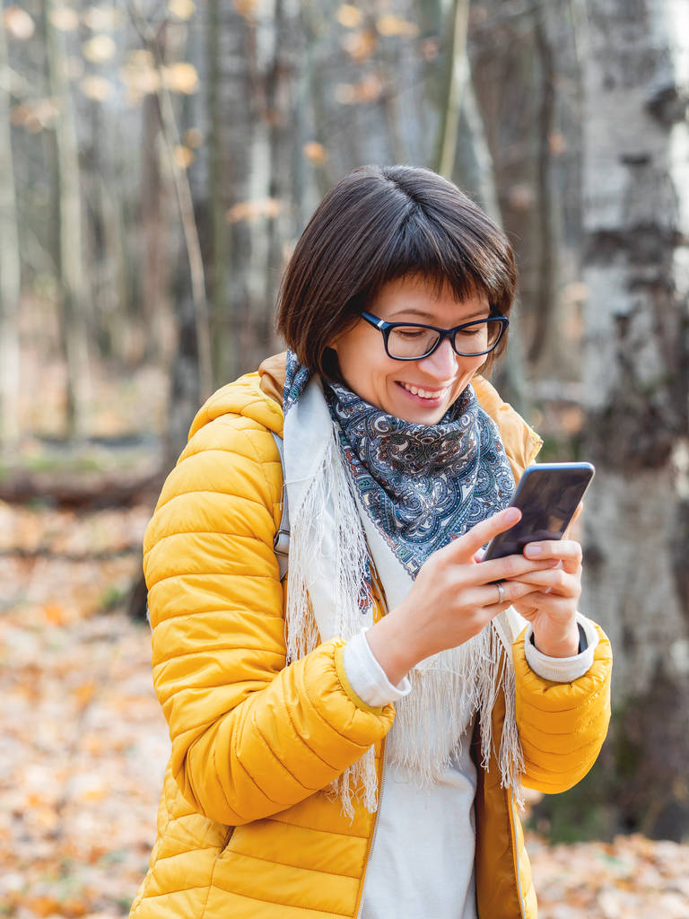 Akıllı telefonlu, parlak sarı ceketli bir kadın. Geniş tebessümlü genç kız sonbahar ormanlarında yürüyor ve akıllı telefondan birine mesaj atıyor.. - Fotoğraf, Görsel
