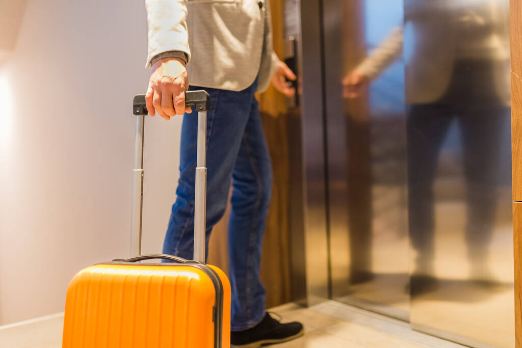 Μοντέρνος ταξιδιώτης άνθρωπος με βαλίτσα πατώντας το σύγχρονο κουμπί ασανσέρ στο λόμπι. - Φωτογραφία, εικόνα