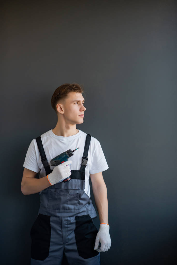 Portrait of a young smiling working man in gray overalls and white t-shirt. Worker holding a electric drill in his hand on a gray background.place for text, copy space - Photo, Image