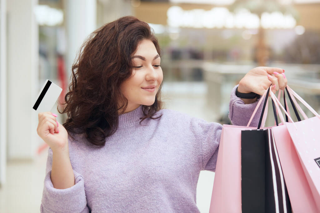 Horizontale shot van gelukkige brunette vrouw met boodschappentassen in de ene hand en creditcard in een andere, kijken naar haar nieuwe kleding, kijkt blij, uiten vreugde, poseren in het winkelcentrum. Levensstijl concept. - Foto, afbeelding