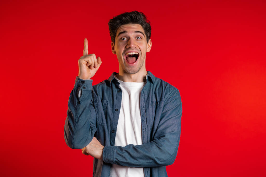 Porträt eines nachdenkenden jungen Mannes mit Idee, der mit erhobenem Finger auf rotem Studiohintergrund zeigt. Lächelnder glücklicher Kerl zeigt Eureka-Geste. - Foto, Bild