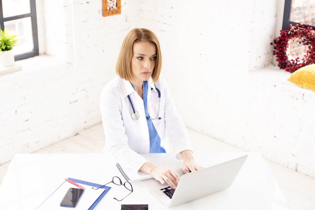 Aufnahme einer Ärztin mittleren Alters, die hinter ihrem Laptop sitzt und arbeitet.  - Foto, Bild