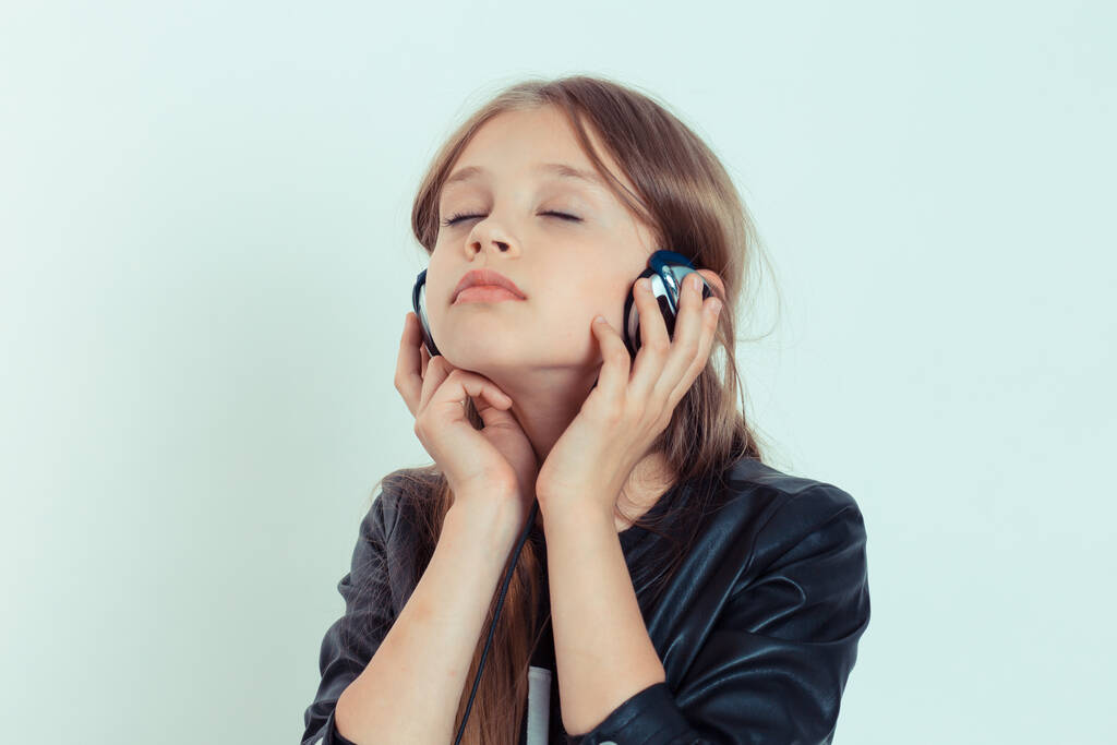 Boldog aranyos kislány élvezi hallgatni a zenét a fülhallgatójában csukott szemmel. Szoros portré fehér gyerek modell fekete bőr kabát elszigetelt fehér kék szürke másolás tér háttér - Fotó, kép