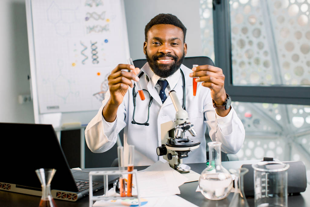 Bellissimo giovane scienziato, chimico, medico africano che indossa un camice da laboratorio e sorride sorridendo sorridendo con due provette con liquido rosso, lavorando in laboratorio. Analisi chimica, concetto di ricerca medica
 - Foto, immagini