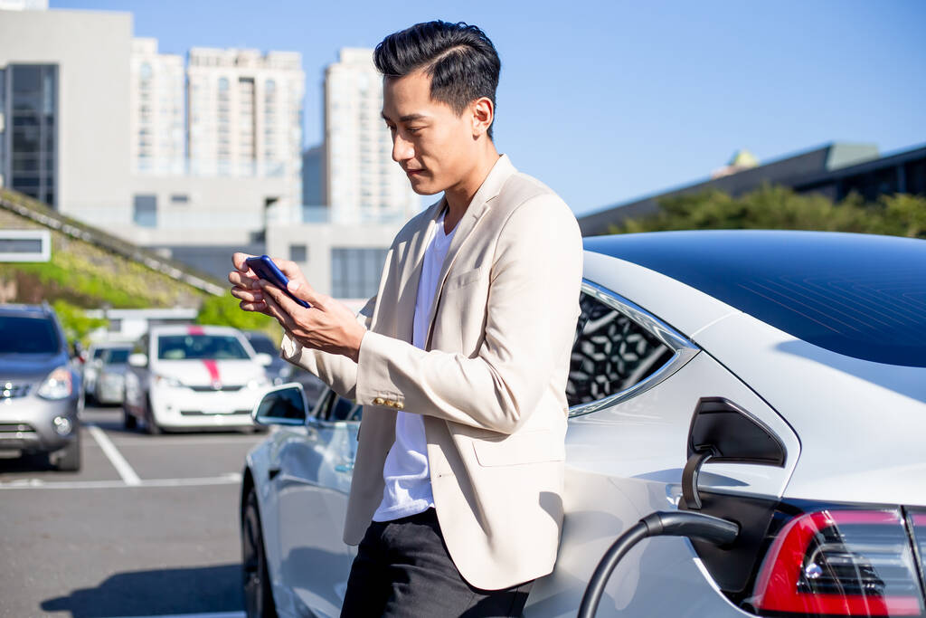 ázsiai férfi használ okos telefon várakozás közben és tápegység csatlakozni elektromos járművek töltésére az akkumulátor az autóban - Fotó, kép