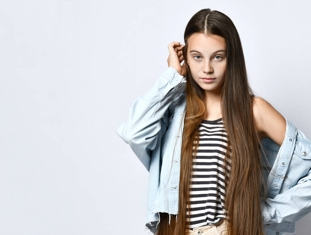 Teenage girl in striped t-shirt, denim jacket and pants. She looking at you, posing isolated on white studio background. Close up - 写真・画像