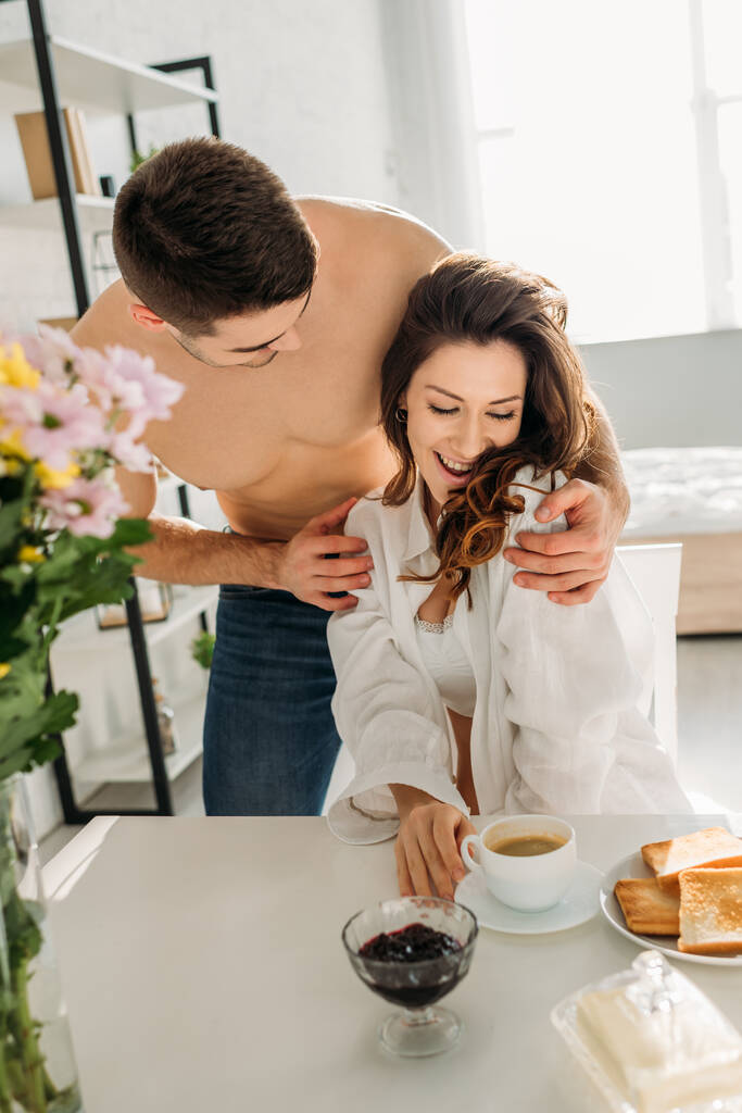 bonito sem camisa homem abraçando feliz menina sentado perto de café da manhã e vaso com flores
 - Foto, Imagem