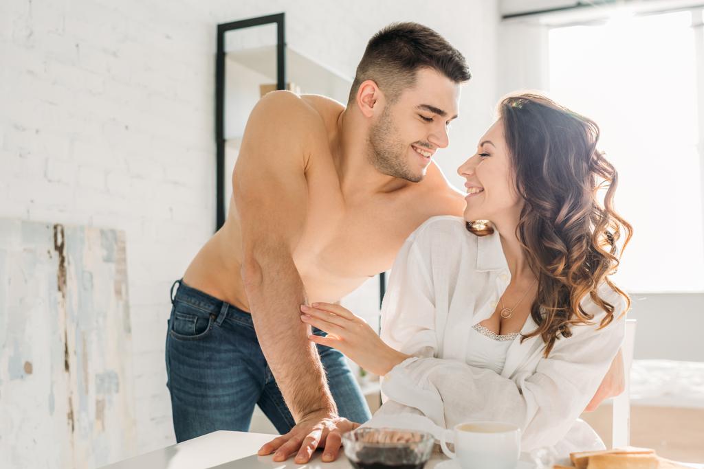 handsome shirtless man looking at happy girl sitting at kitchen table in white shirt - Photo, Image