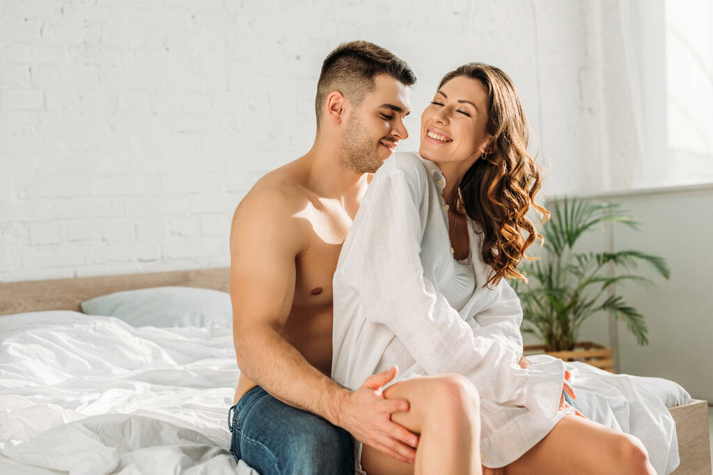 glimlachende shirtloze man aanraken been van sexy, vrolijk meisje terwijl zitten op bed - Foto, afbeelding