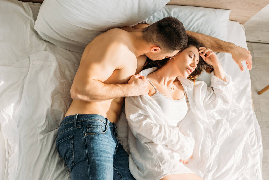 top view of shirless man in jeans przytulanie dziewczyna leży w łóżku z zamkniętymi oczami - Zdjęcie, obraz