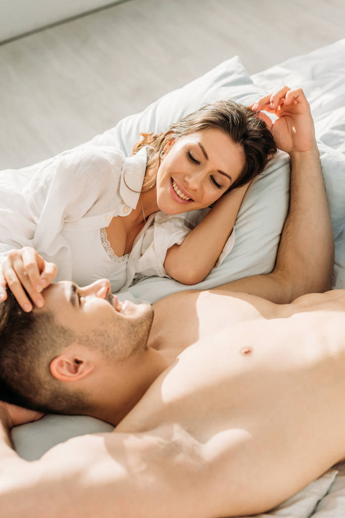 happy, sensual girl and shirtless man touching heads of each other while lying in bed together - Photo, Image