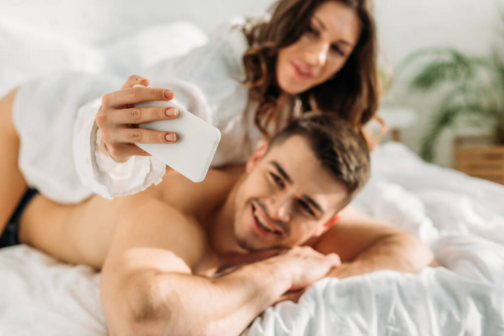 επιλεκτική εστίαση χαμογελαστό κορίτσι λήψη selfie στο smartphone με σέξι χαρούμενο άνδρα, ενώ βρίσκονται στο κρεβάτι μαζί - Φωτογραφία, εικόνα