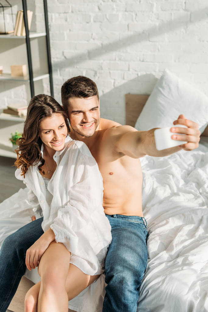 selective focus of cheerful man taking selfie on smartphone with sexy girlfriend in bedroom - Photo, Image