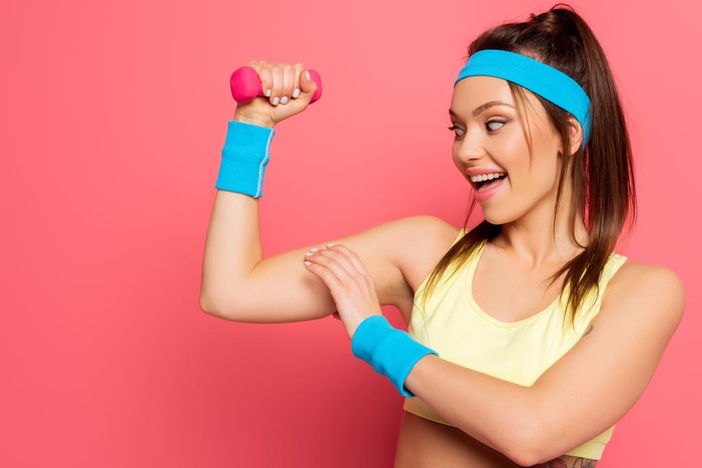 opgewonden sportvrouw aanraken biceps tijdens de training met halter op roze achtergrond - Foto, afbeelding