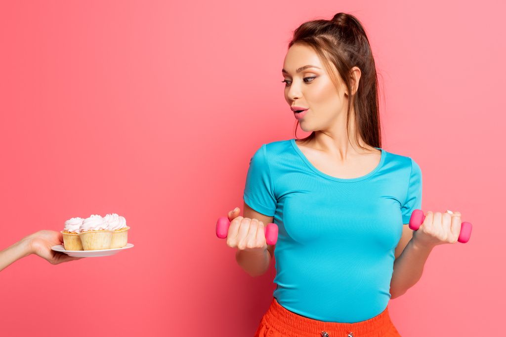 surprised sportswoman exercising with dumbbells near female hand with plate of delicious cupcakes on pink background - Photo, Image