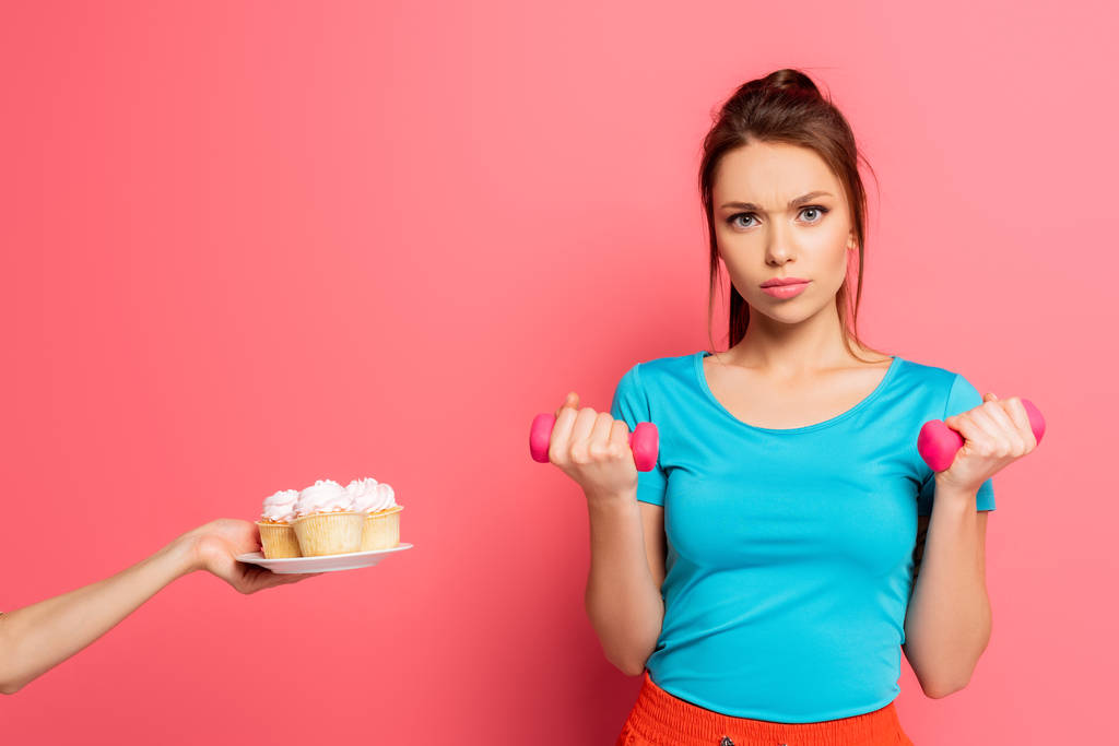 confident sportswoman exercising with dumbbells near female hand with plate of delicious cupcakes on pink background - Photo, Image