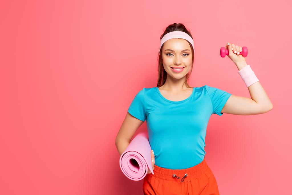 gelukkig sportvrouw met halter en fitness mat terwijl glimlachen op de camera op roze achtergrond - Foto, afbeelding