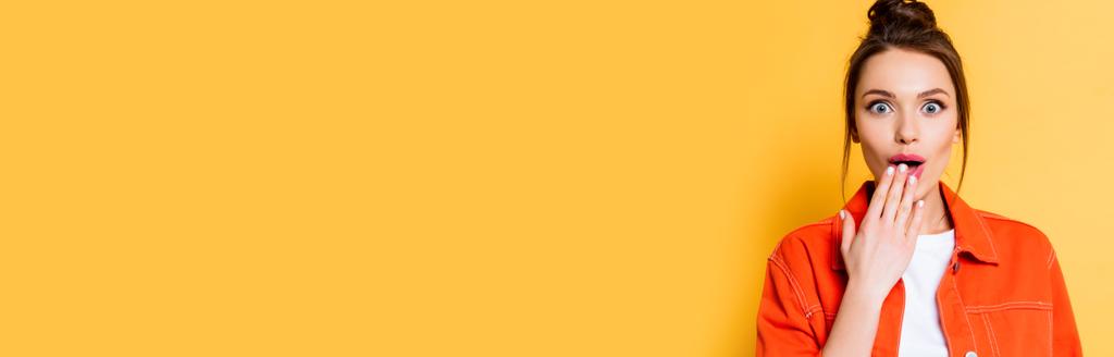 panoramic shot of shocked girl looking at camera while covering mouth with hand on yellow background - Photo, Image