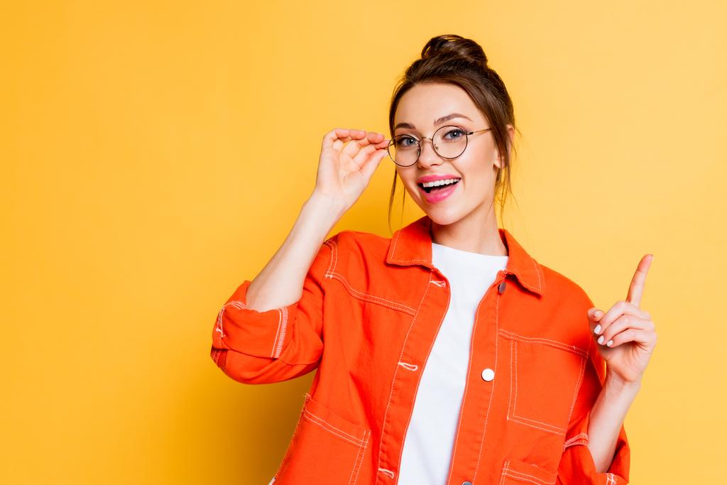 happy student touching eyeglasses and showing idea gesture while looking at camera on yellow background - Photo, Image
