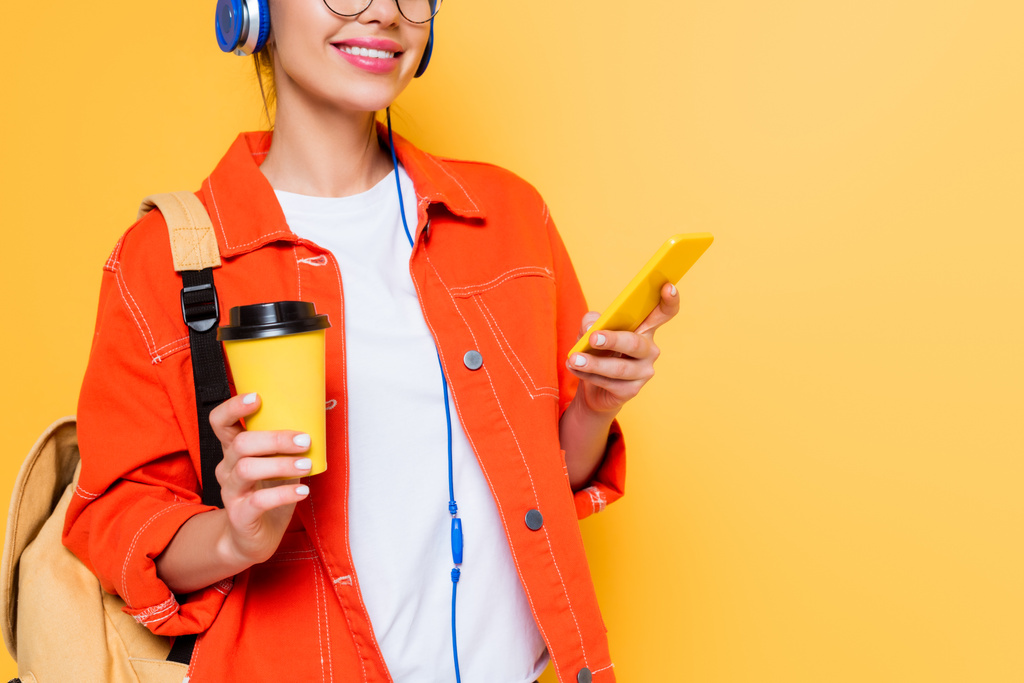 περικοπή άποψη του μαθητή σε ακουστικά chatting στο smartphone, ενώ κρατώντας καφέ για να πάει απομονωθεί σε κίτρινο - Φωτογραφία, εικόνα