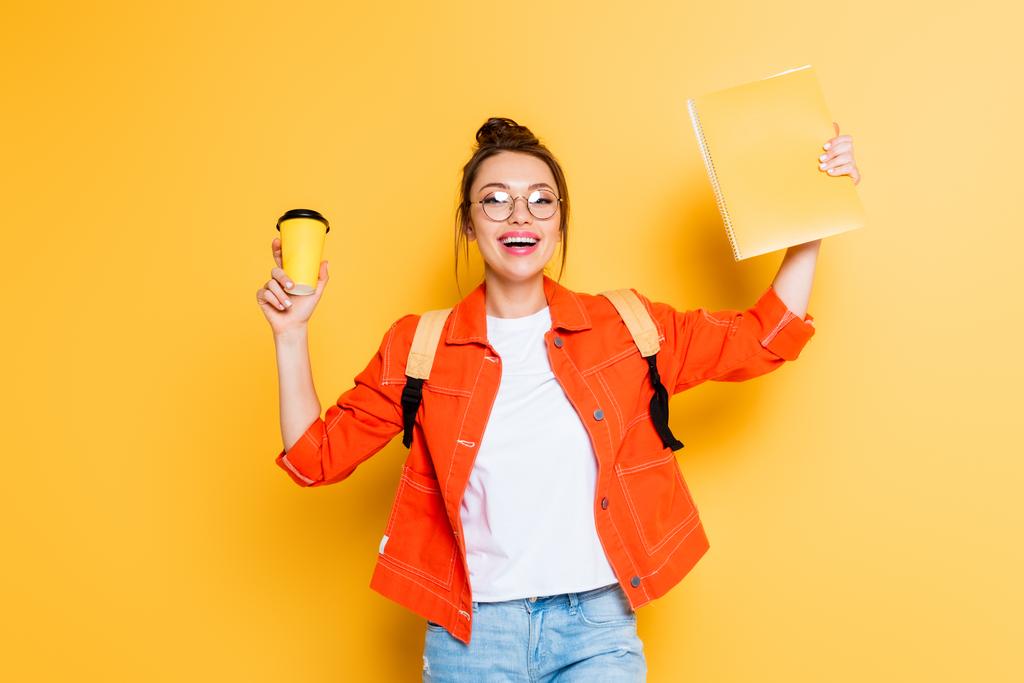 excited student holding coffee to go and copy book in raised hands while smiling at camera on yellow background - Photo, Image
