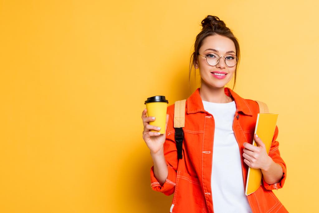 ελκυστική, ευτυχής φοιτητής κρατώντας καφέ για να πάει και σημειωματάριο, ενώ χαμογελά στην κάμερα σε κίτρινο φόντο - Φωτογραφία, εικόνα
