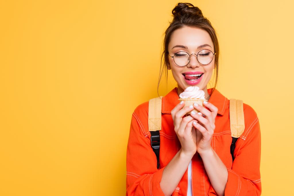 estudante feliz com olhos fechados colando os lábios enquanto segurando delicioso cupcake no fundo amarelo
 - Foto, Imagem