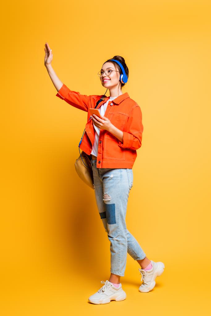 Ganzkörperansicht des fröhlichen Studenten mit Kopfhörern, der mit der Hand wedelt, während er auf gelbem Hintergrund wegschaut - Foto, Bild