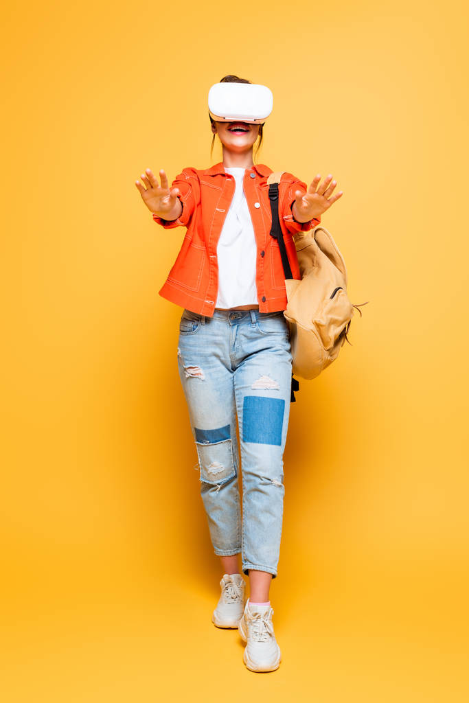 взволнованная девушка с рюкзаком, стоящая с протянутыми руками, используя гарнитуру VR на желтом фоне
 - Фото, изображение