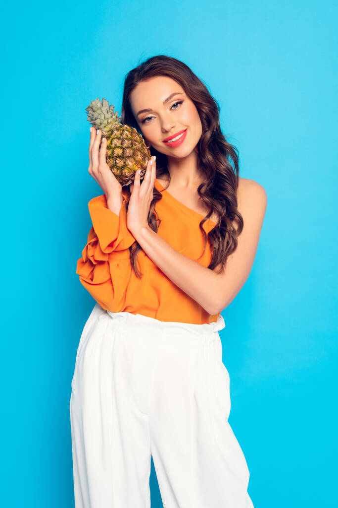 atrakcyjna, uśmiechnięta dziewczyna patrząc w kamerę trzymając dojrzały ananas odizolowany na niebiesko - Zdjęcie, obraz