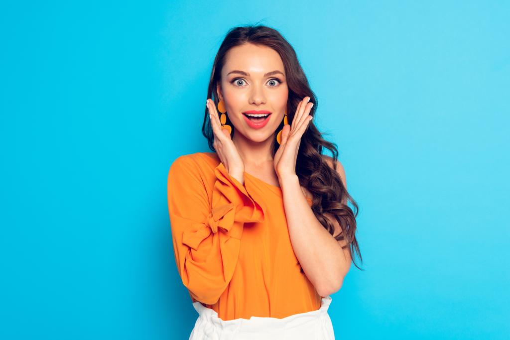 excited stylish girl holding hands near face and looking at camera on blue background - Photo, Image