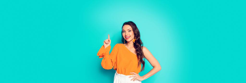 panoramic shot of cheerful, stylish girl showing idea gesture while standing with hand on hip on turquoise background - Photo, Image
