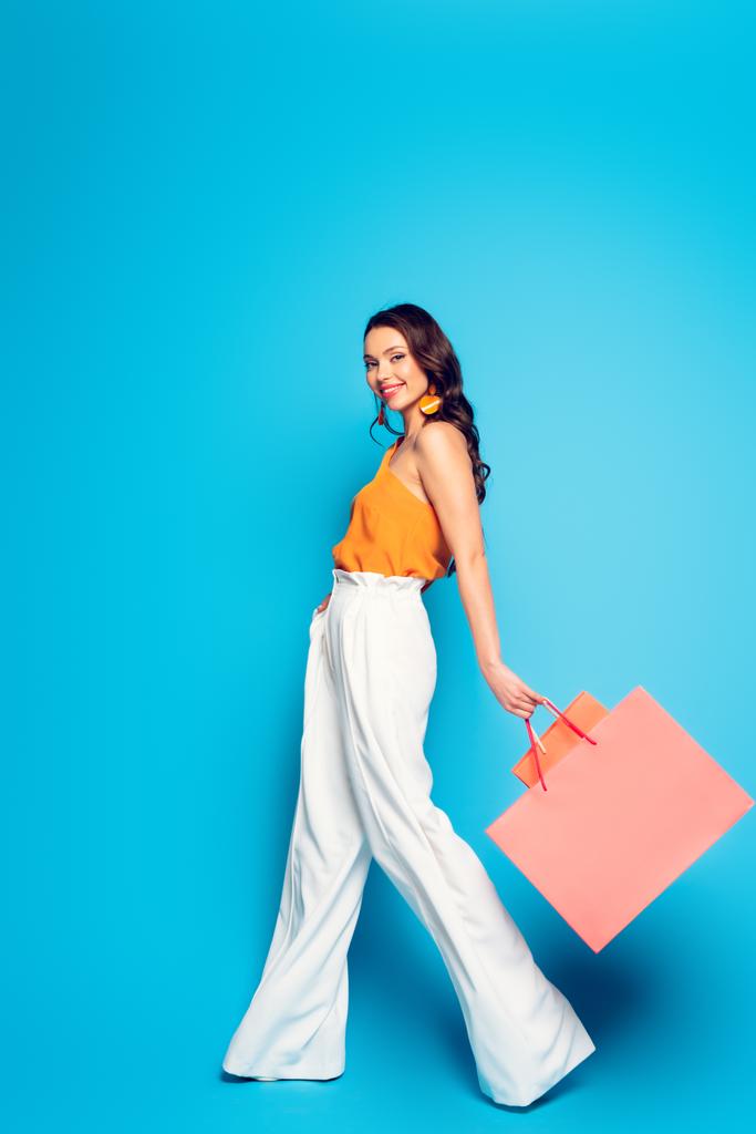 volledige weergave van vrolijke stijlvolle vrouw wandelen met roze boodschappentassen en glimlachen op camera op blauwe achtergrond - Foto, afbeelding