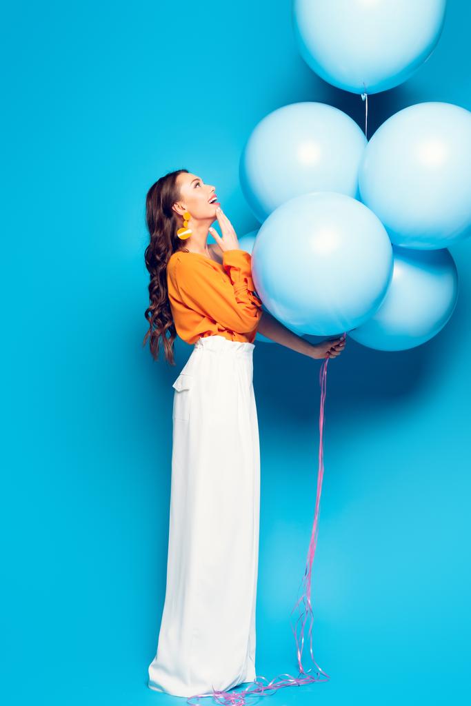 užaslá stylová žena dotýkající se brady při pohledu na velké slavnostní balónky na modrém pozadí - Fotografie, Obrázek