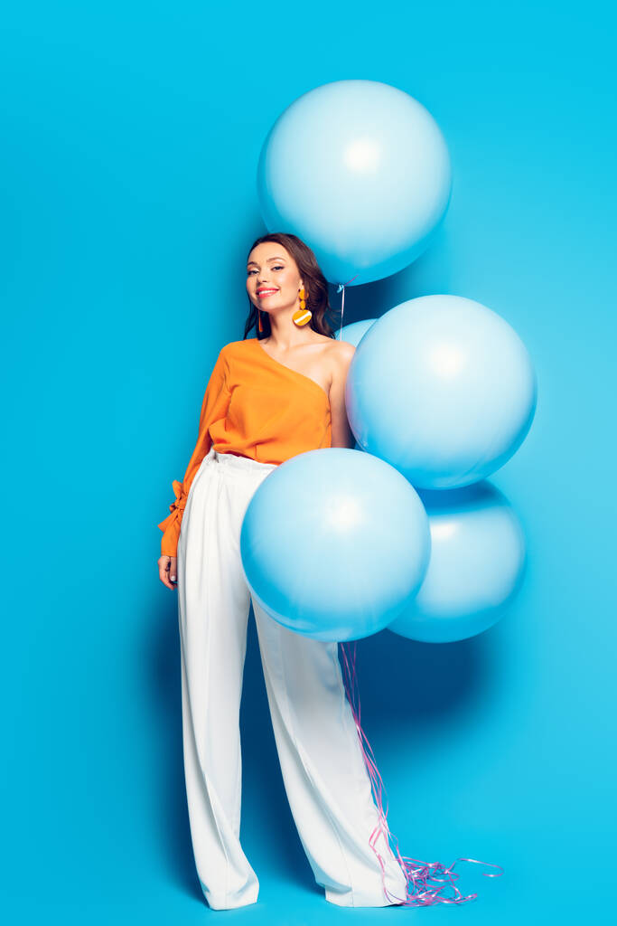 full length view of cheerful stylish woman with big festive balloons smiling at camera on blue background - Photo, Image