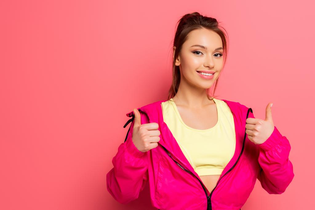 fiatal vidám sportasszony mutatja hüvelykujját felfelé, miközben mosolygott a kamera rózsaszín háttér - Fotó, kép
