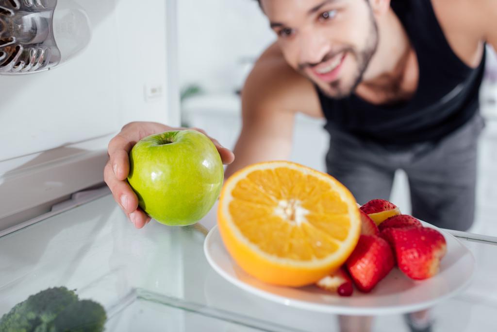 selectieve focus van een gelukkig man die appel uit de koelkast haalt  - Foto, afbeelding