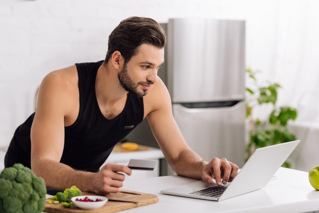 όμορφος άντρας που κρατά πιστωτική κάρτα ενώ χρησιμοποιεί το laptop στην κουζίνα  - Φωτογραφία, εικόνα