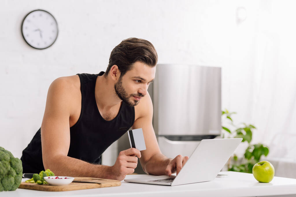 handsome man holding credit card while using laptop near apple and broccoli  - Photo, Image