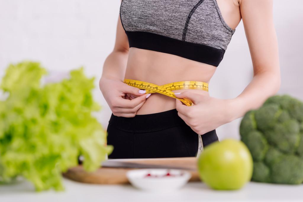 cropped view of sportive girl measuring waist near fresh food  - Photo, Image